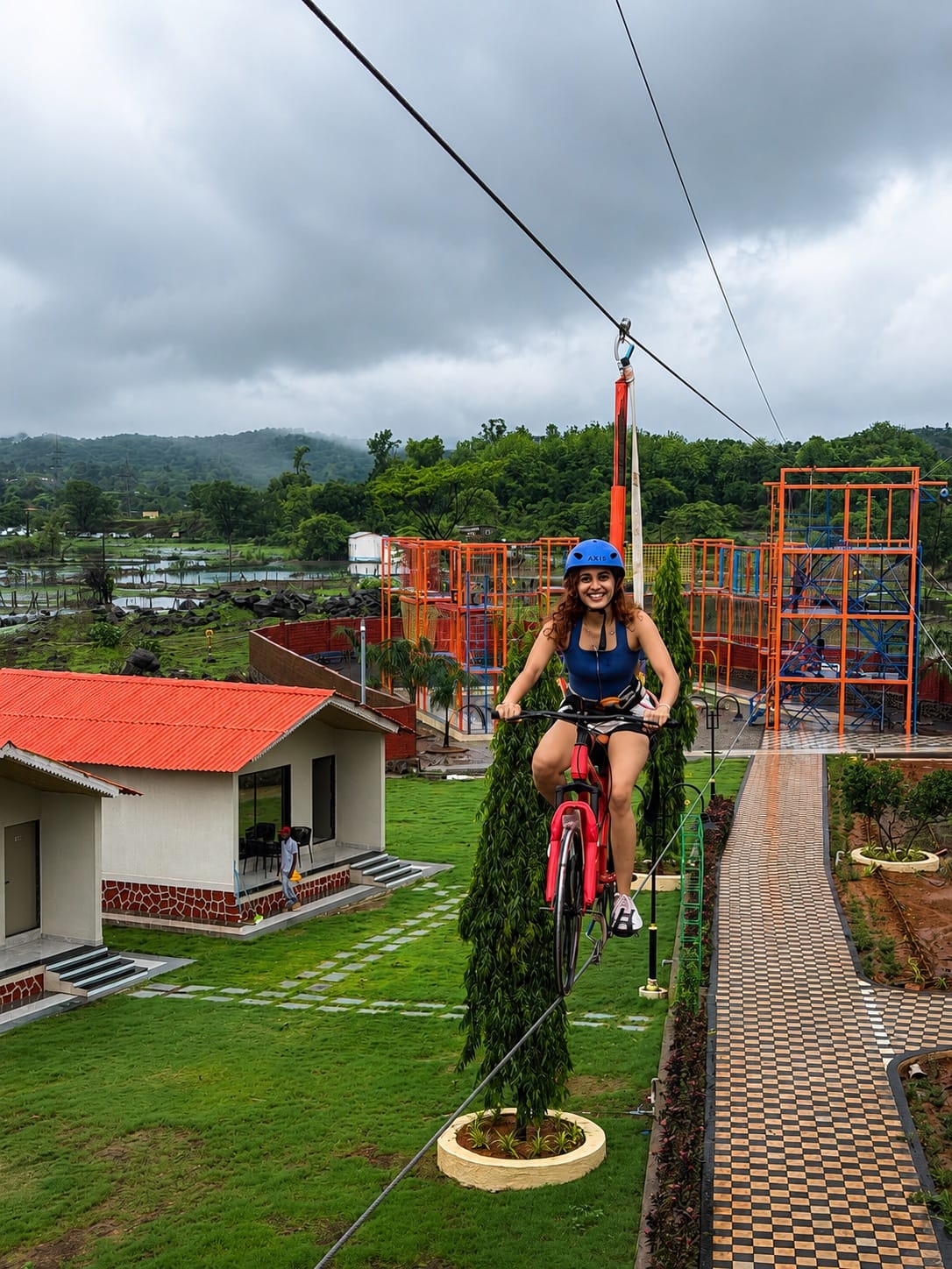 A guest pedalling the sky cycle along its cable above the cottages, with the orange climbing tower beyond