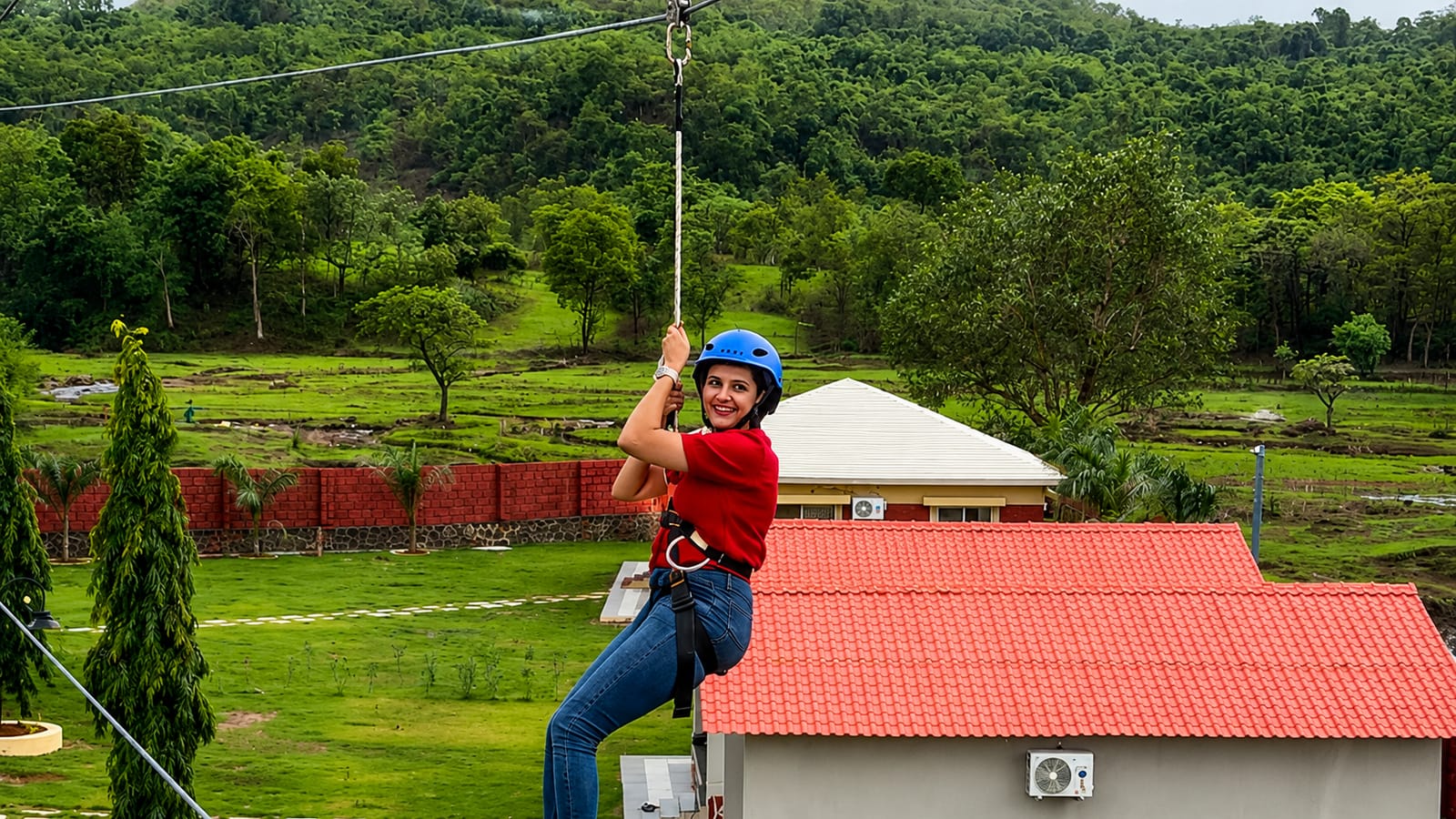 A harnessed guest riding the zipline over the farm, with the red-roofed cottages and green hills behind